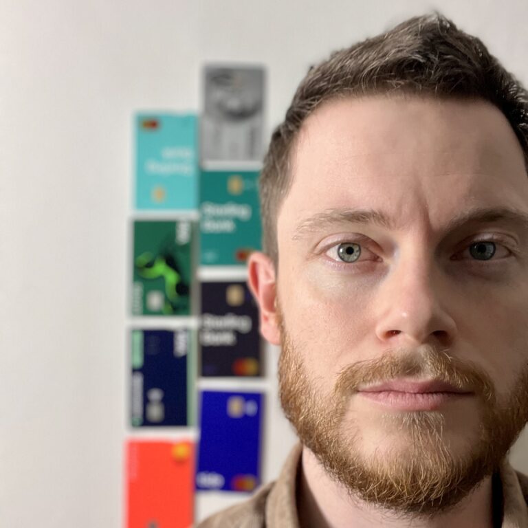 Joe - a white man with short dark hair and a beard - is in crisp focus in the right of the frame. A line of colourful credit and debit cards, out of focus, line the wall behind him.